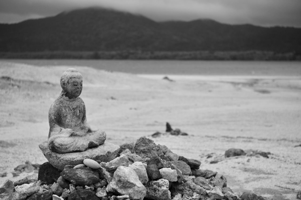 Black and white photo of Buddha statue on rocky beach in Mutsu, Japan.